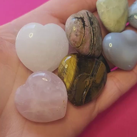 Close-up video of a hand holding several medium polished mystery crystal heart stones against a bright pink background, showcasing their smooth surface, varied colors, and natural crystal patterns — ideal for crystal collections, meditation, healing, and gifting.