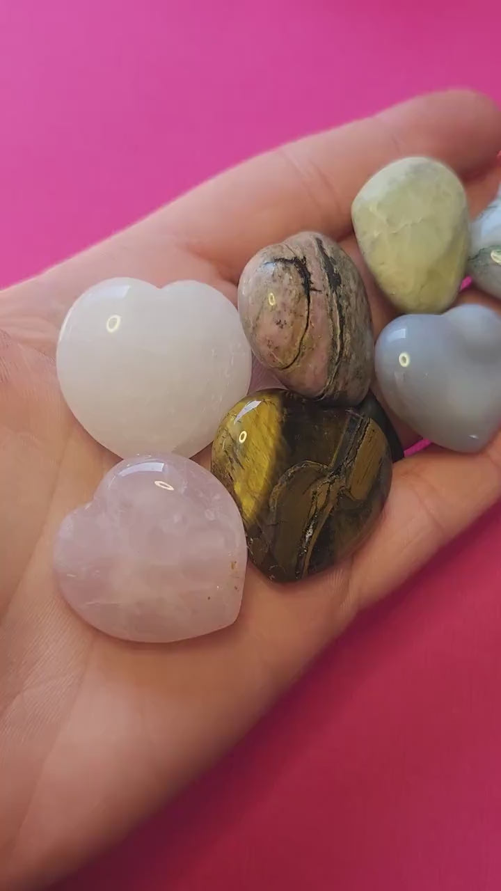 Close-up video of a hand holding several medium polished mystery crystal heart stones against a bright pink background, showcasing their smooth surface, varied colors, and natural crystal patterns — ideal for crystal collections, meditation, healing, and gifting.