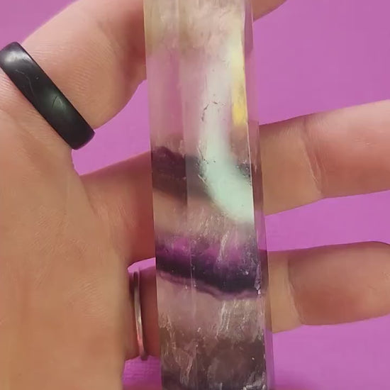 Video of a polished Fluorite crystal tower held in hand against a purple background, showcasing natural green, purple, and clear banding, polished gemstone point for mental clarity, meditation, chakra balancing, energy cleansing, spiritual healing, and metaphysical crystal décor