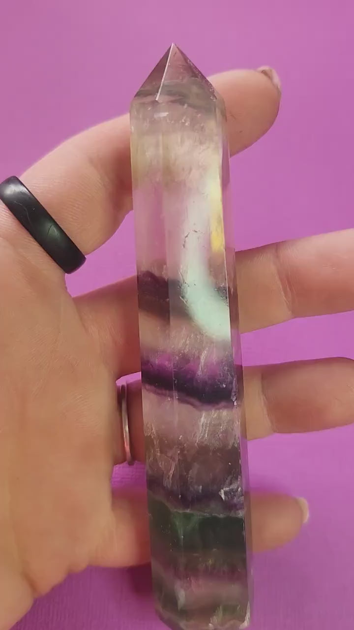 Video of a polished Fluorite crystal tower held in hand against a purple background, showcasing natural green, purple, and clear banding, polished gemstone point for mental clarity, meditation, chakra balancing, energy cleansing, spiritual healing, and metaphysical crystal décor