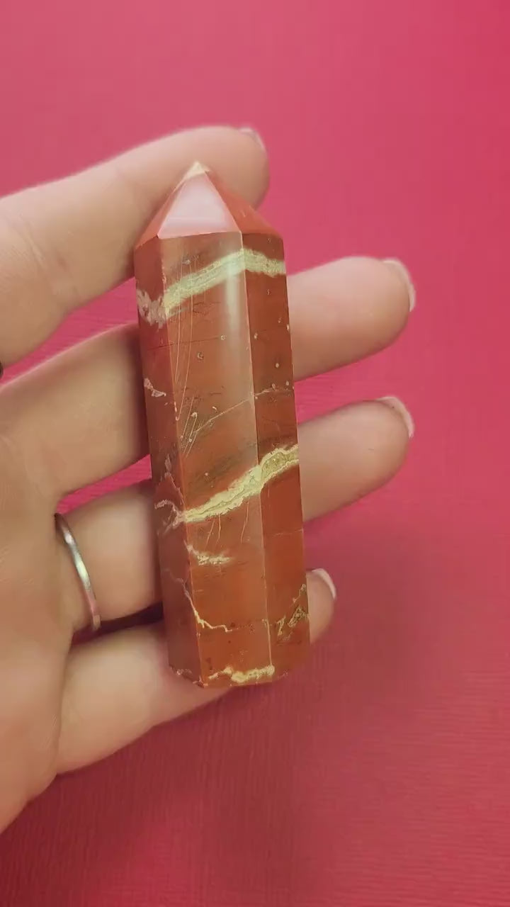 Video of a hand holding a polished Red Jasper crystal tower up close against a red background, highlighting its deep red color, natural striations, and smooth polished finish. Perfect for crystal healing, grounding, energy work, meditation, Reiki, and spiritual decor. Ideal for crystal collectors, home altars, and intention setting.