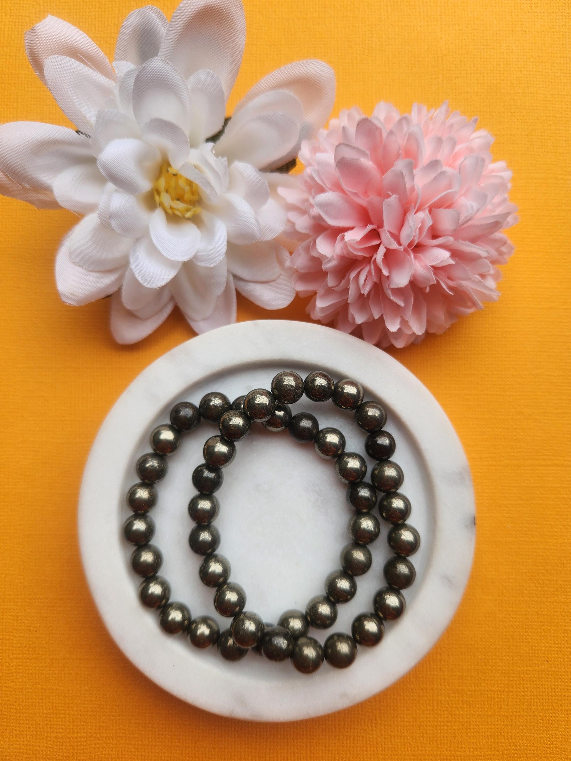 Two pyrite 8mm beaded bracelets displayed in a white dish on an orange background with flower props featuring metallic gold crystal beads for abundance confidence protection energy manifestation jewelry gift