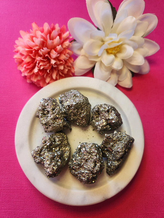 Raw Pyrite crystal pieces displayed in a white dish with two flower props on a bright pink background. The metallic gold stones shimmer beautifully. Perfect for abundance, manifestation, and crystal decor