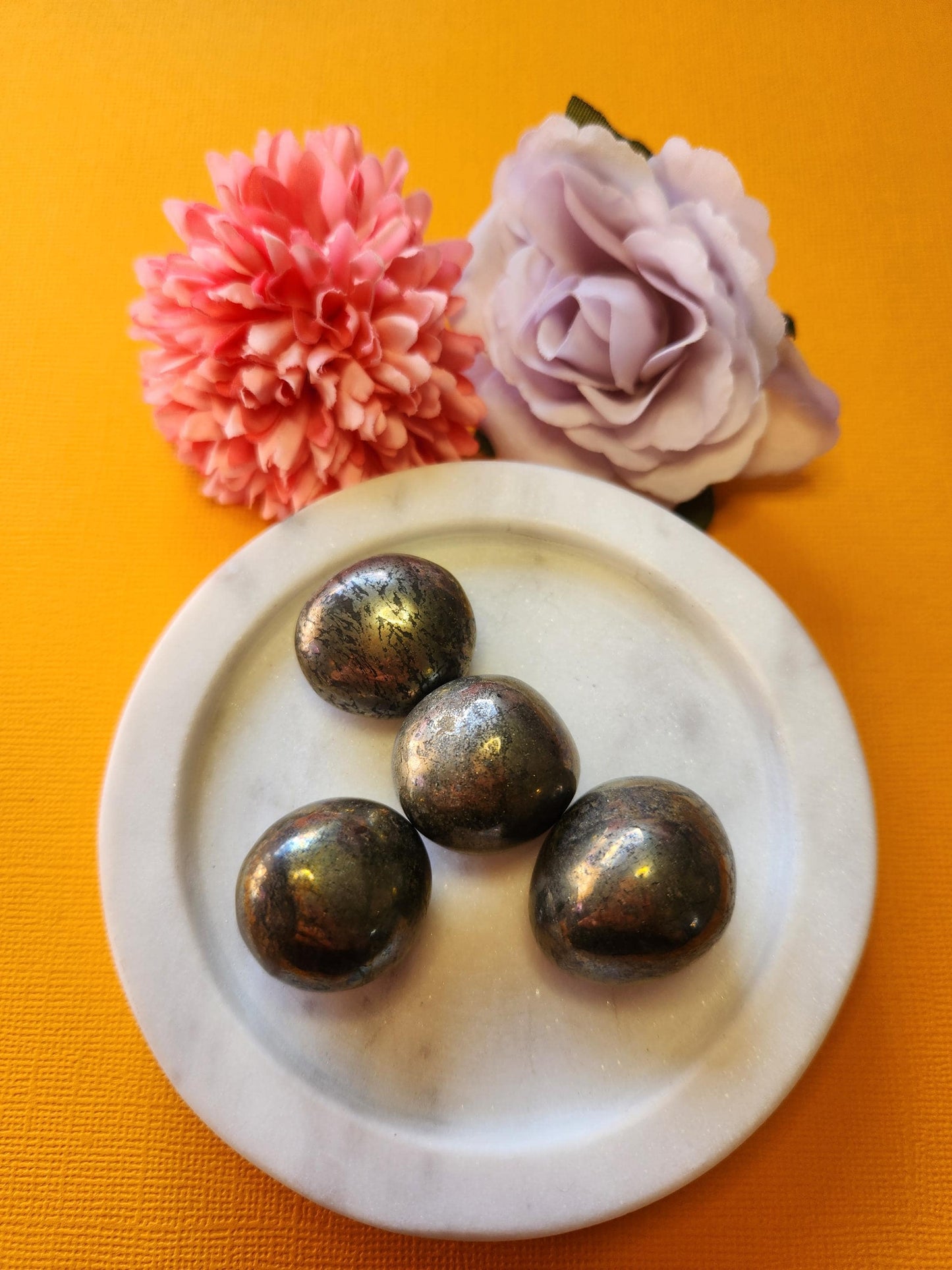 Pyrite tumble stones in a white dish with two flower props on a yellow background, polished golden crystals for abundance, confidence, and protection.