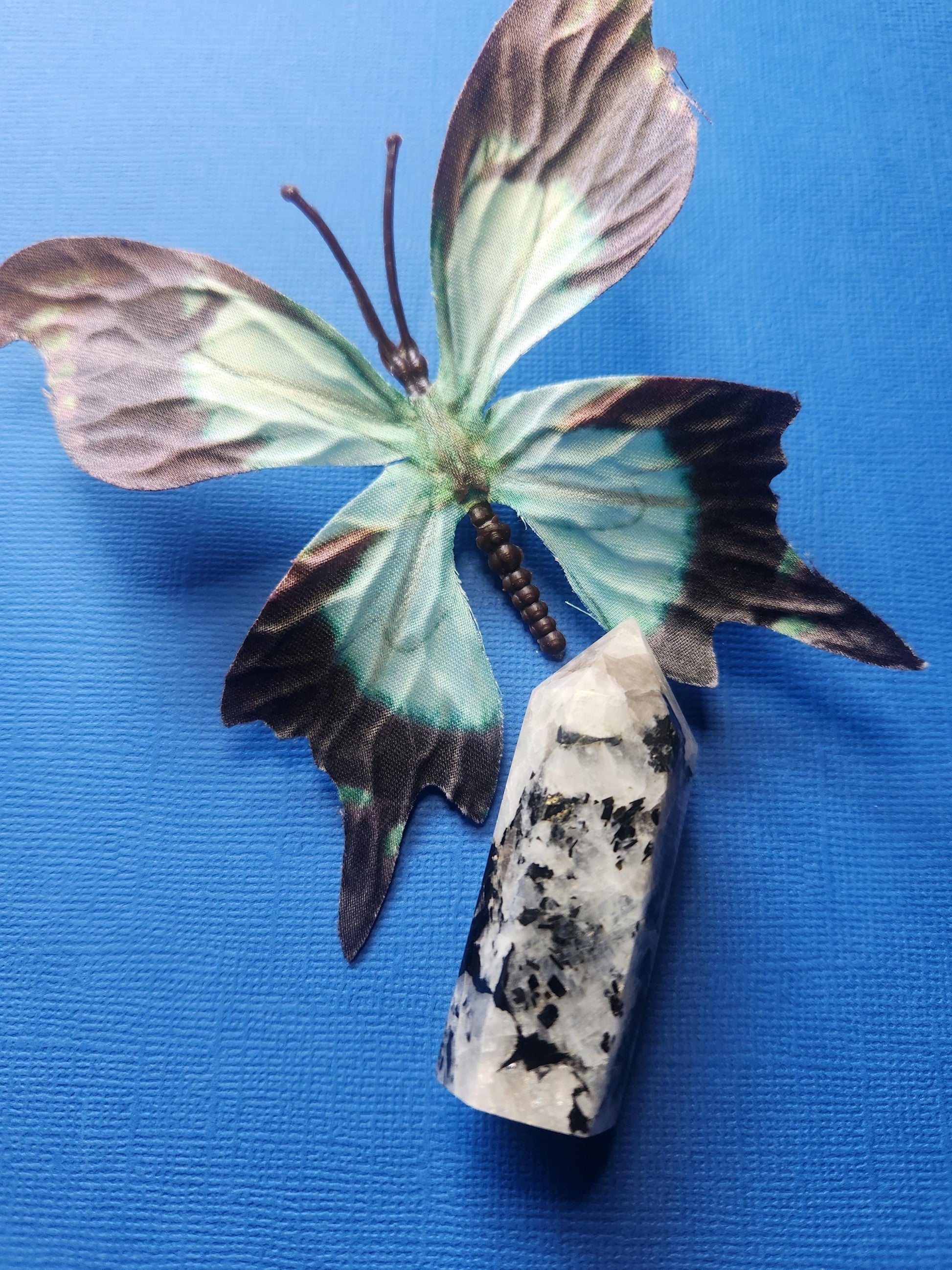 Rainbow moonstone crystal tower displayed with a blue butterfly prop, set against a dark blue background. Hand-carved and polished, this tower promotes intuition, emotional balance, and spiritual growth — perfect for meditation, crystal collections, and home décor.