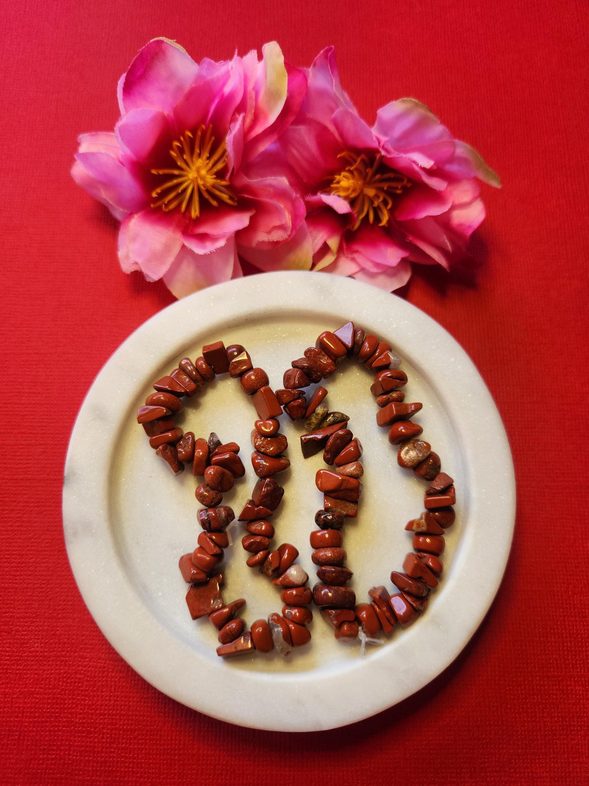 Two Red Jasper chip bracelets displayed in a white dish with two flower props on a red background, featuring polished deep red crystal chips that promote grounding, strength, and stability — perfect Root Chakra healing crystal jewelry and protective energy gift.