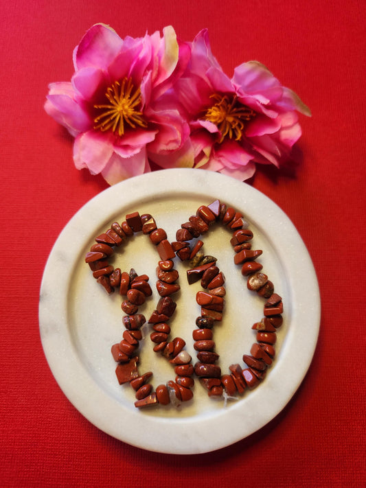 Two Red Jasper chip bracelets displayed in a white dish with two flower props on a red background, featuring polished deep red crystal chips that promote grounding, strength, and stability — perfect Root Chakra healing crystal jewelry and protective energy gift.