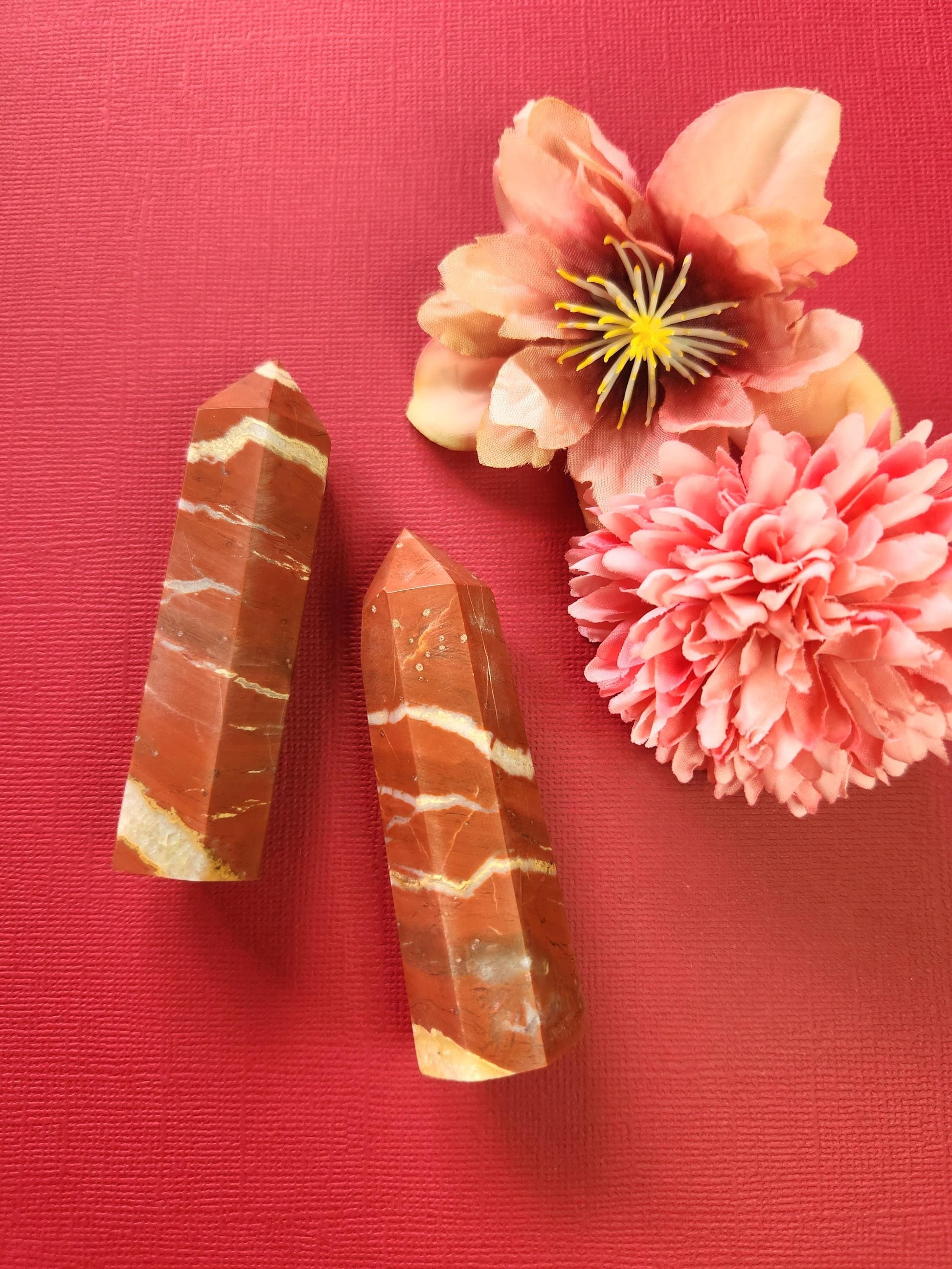 Two polished Red Jasper crystal towers displayed on a red background with two decorative flower props. The vibrant red tones of the crystals stand out, showcasing their grounding energy and natural patterns. Ideal for crystal lovers, spiritual decor, and meditation photography.