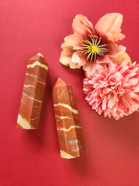 Two polished Red Jasper crystal towers displayed on a red background with two decorative flower props. The vibrant red tones of the crystals stand out, showcasing their grounding energy and natural patterns. Ideal for crystal lovers, spiritual decor, and meditation photography.
