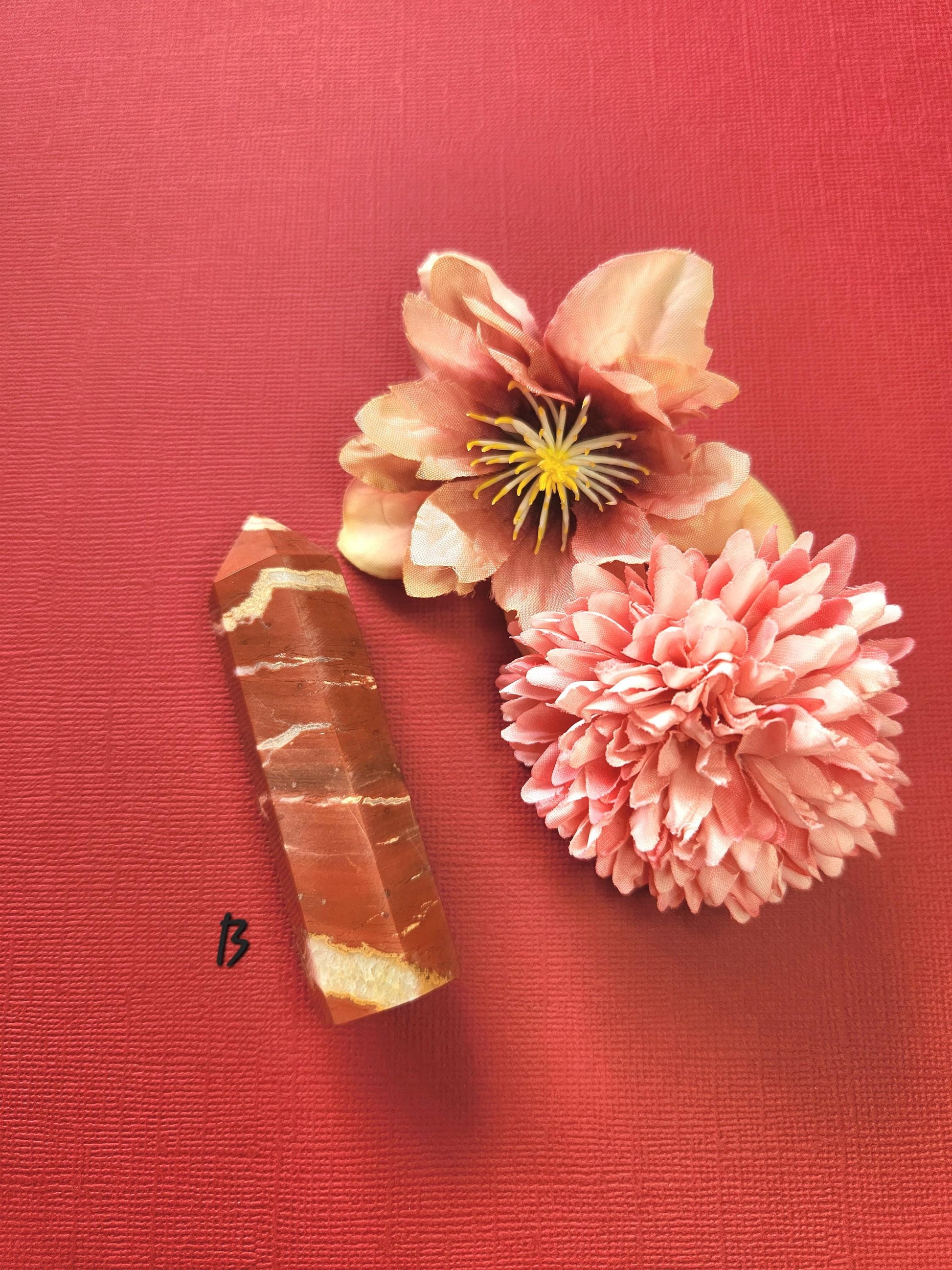 Polished Red Jasper crystal tower (Tower B) displayed on a red background with two decorative flower props, highlighting its rich red tones, natural striations, and grounding energy. Ideal for meditation, Reiki, crystal healing, spiritual decor, home altars, and crystal collectors.