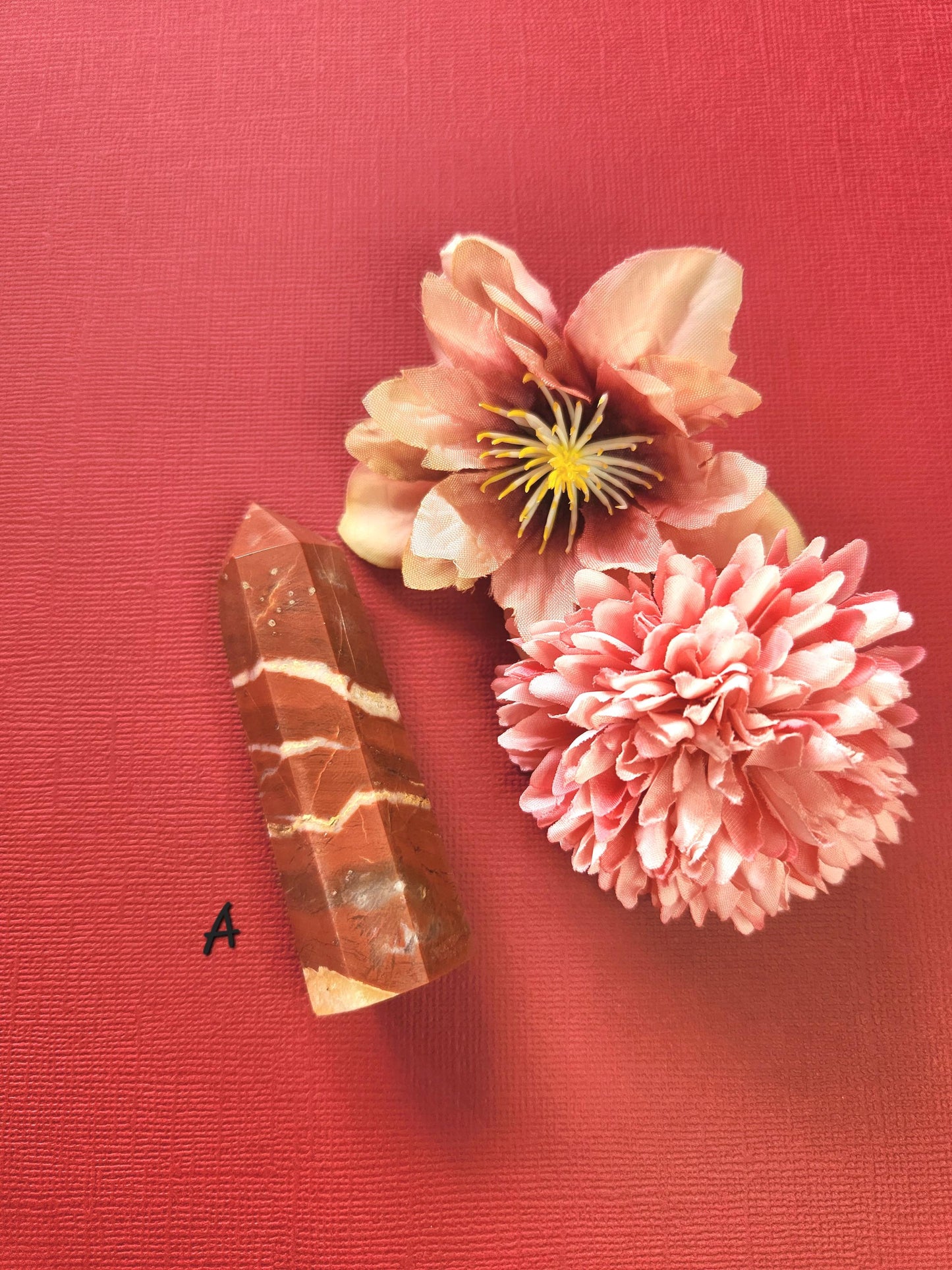 Polished Red Jasper crystal tower (Tower A) displayed on a red background with two decorative flower props, showing its deep red color, natural patterns, and grounding energy. Perfect for meditation, Reiki, crystal healing, spiritual decor, and home altars.
