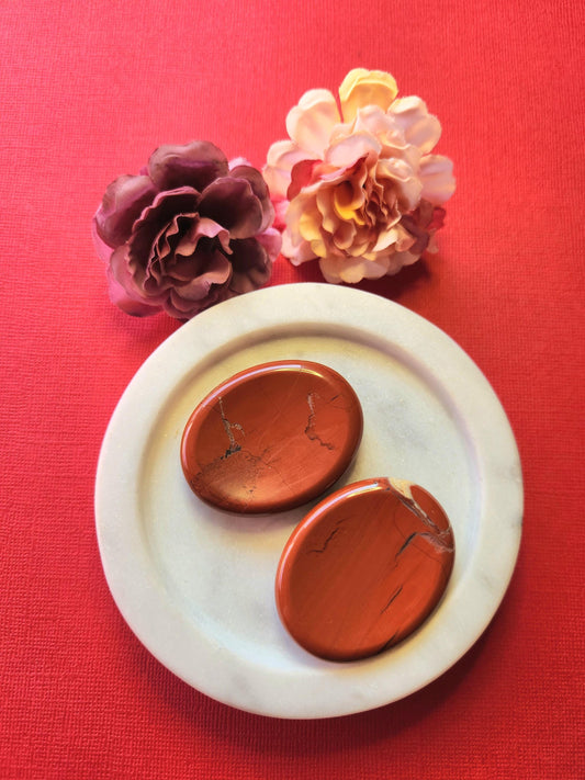 Two Red Jasper worry stones displayed on a white dish with two flower props, set against a red background. Vibrant red thumb stones for grounding energy, vitality, and emotional strength.