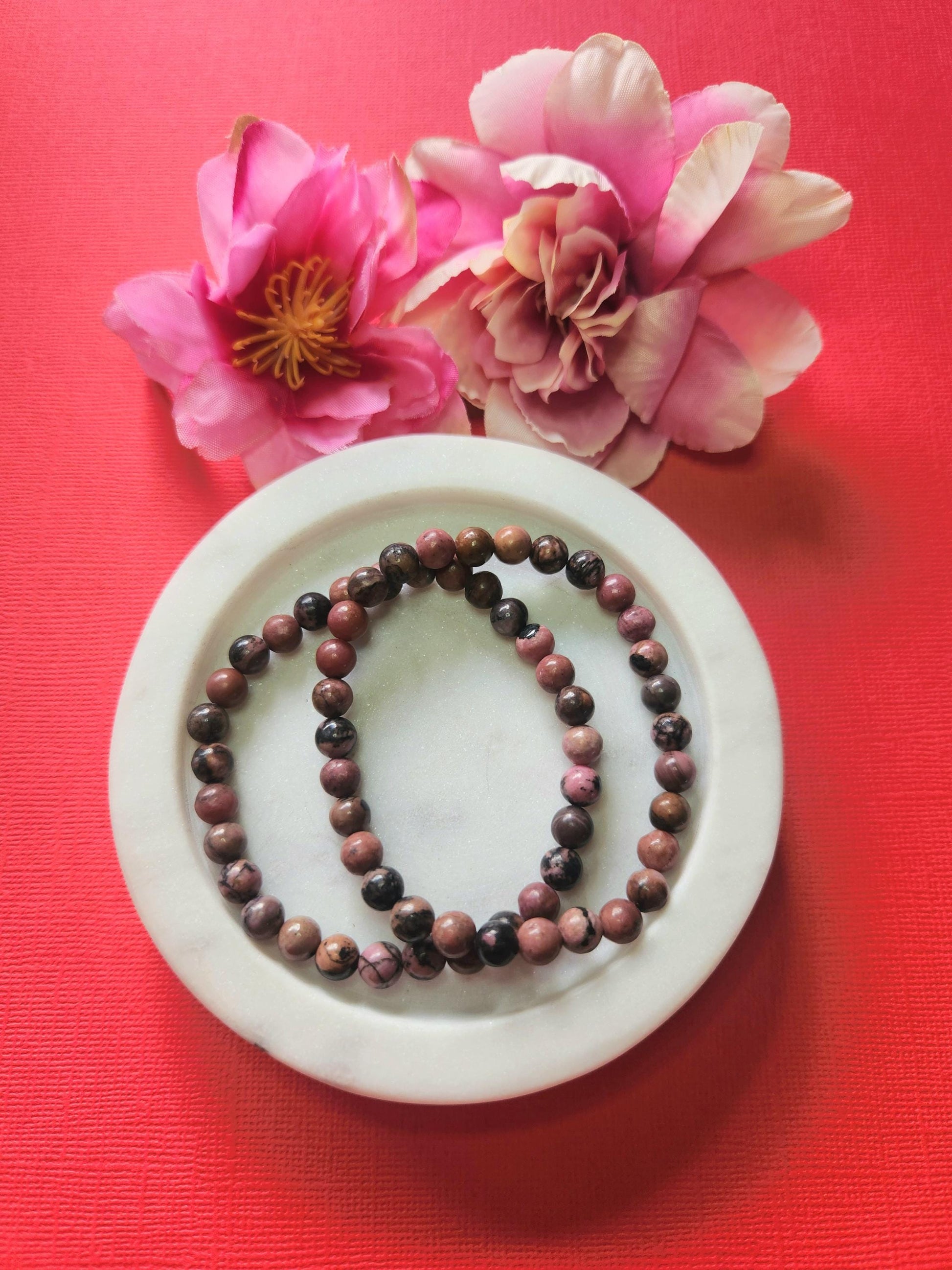 Rhodonite 6mm beaded crystal bracelets in a white dish with flower props on a red background, featuring natural pink and black gemstone beads for love, emotional healing, and self-compassion energy jewelry photography.