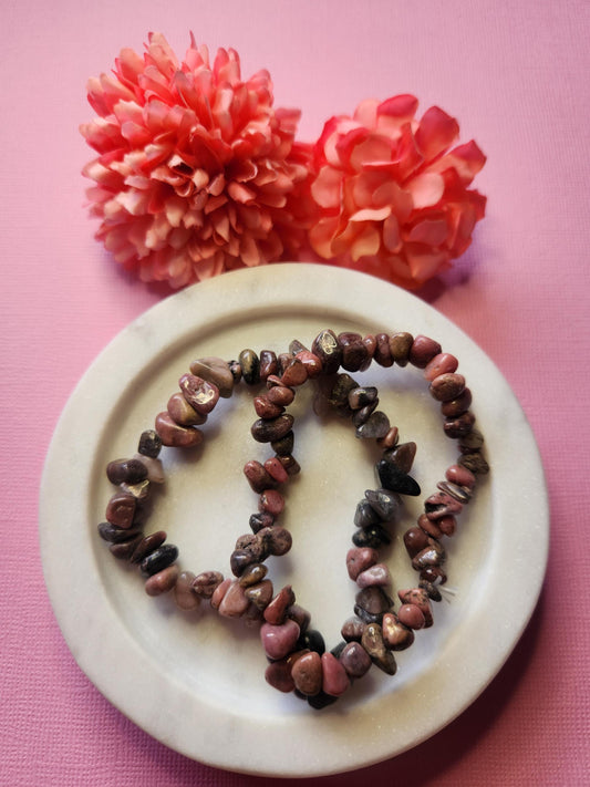 Two Rhodonite chip bracelets displayed in a white dish with two flower props on a light pink background. Natural gemstone jewelry for heart healing, emotional balance, chakra work, meditation, and crystal lovers. Perfect gift for her, self-love accessory, and spiritual bracelet.
