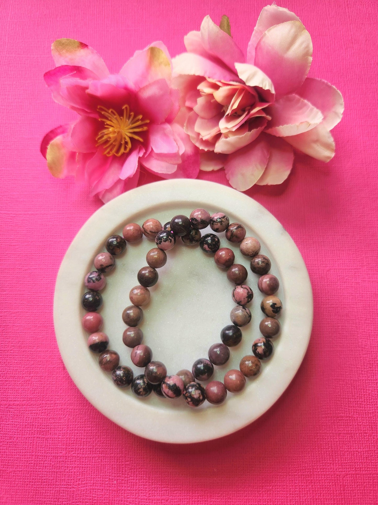 Two Rhodonite 8mm beaded bracelets in white dish with two flower props on pink background, natural rhodonite gemstone beads, handmade crystal jewellery, emotional healing and love energy stone bracelet product photo