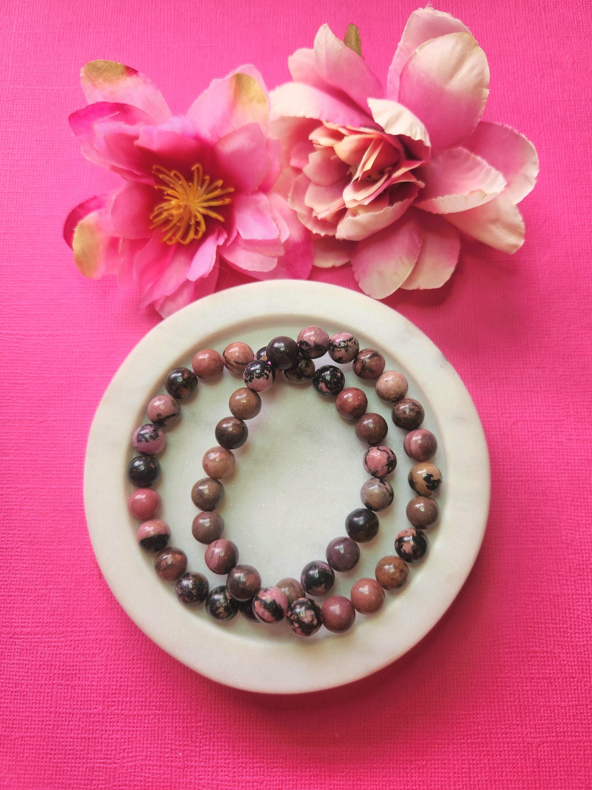 Two Rhodonite 8mm beaded bracelets in white dish with two flower props on pink background, natural rhodonite gemstone beads, handmade crystal jewellery, emotional healing and love energy stone bracelet product photo