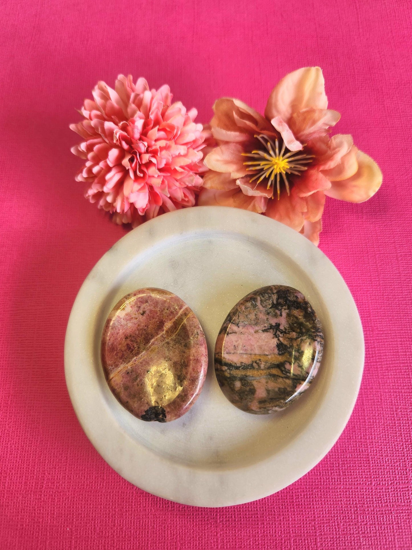 Rhodonite worry stone displayed in a white dish on a pink background with soft flower props featuring polished pink and black crystal stone for emotional healing stress relief grounding calming energy and crystal decor