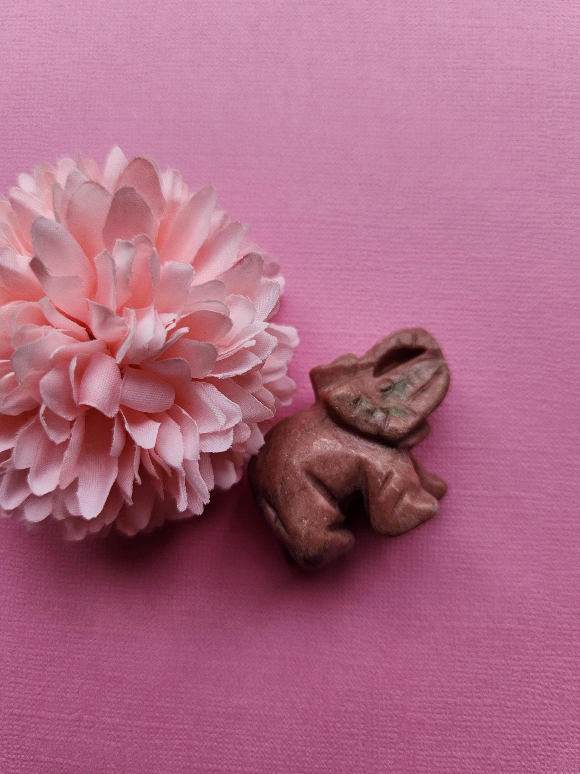 Photo of a Rhodonite crystal carving displayed on a light pink background with one flower prop, showing smooth pink and black stone details — loving crystal decor for emotional healing, self-love, and heart chakra energy