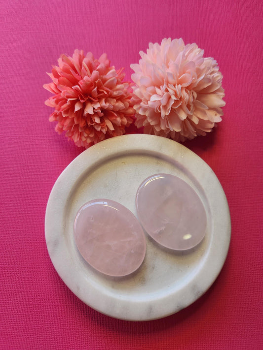 Two Rose Quartz worry stones displayed on a white dish with two flower props, set against a bright pink background. Soft pink thumb stones for love, compassion, emotional healing, and heart chakra energy.