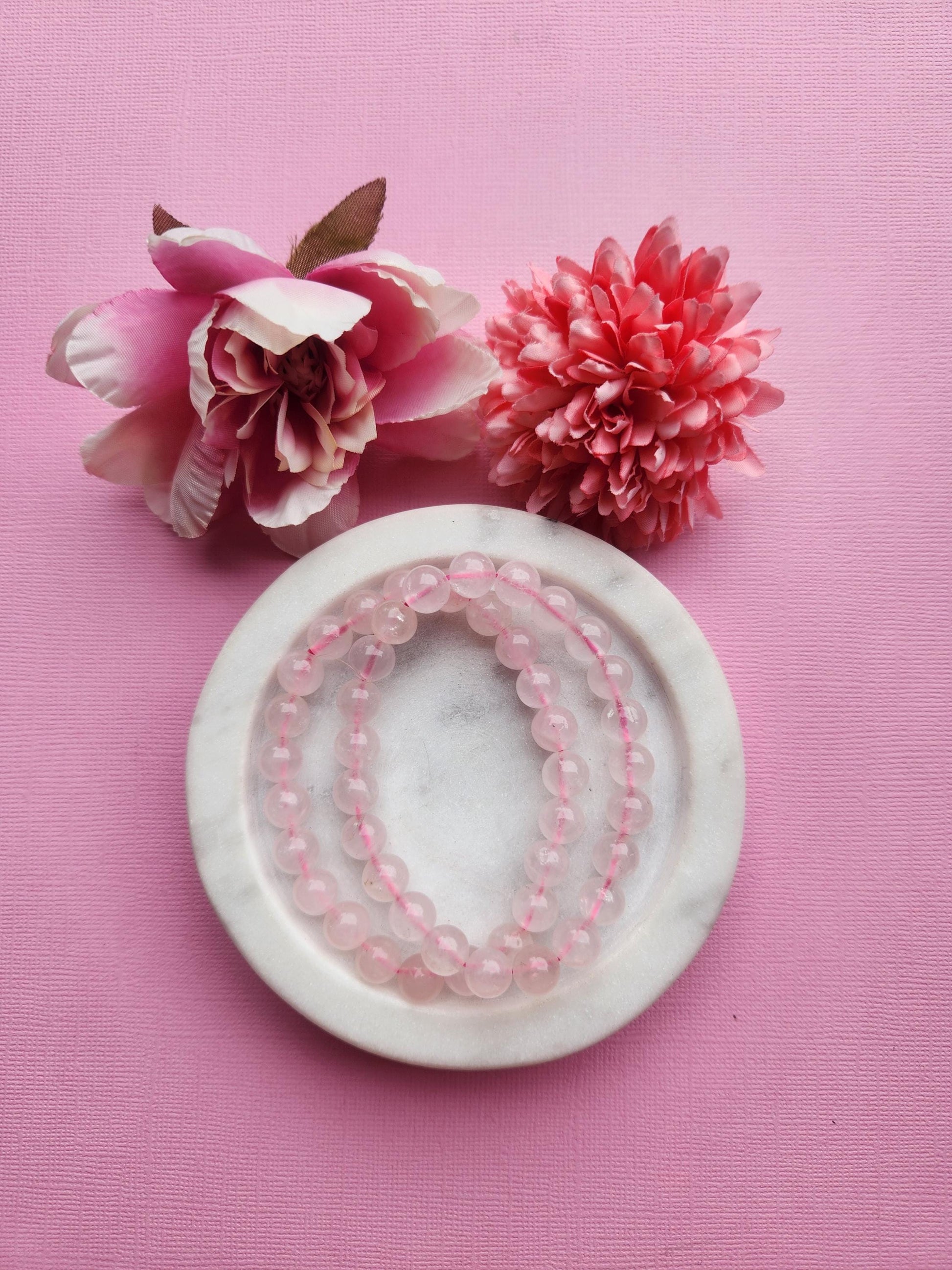 Two rose quartz 8mm beaded bracelets displayed in a white dish on a pink background with soft flower props featuring polished pink crystal beads for love heart healing emotional balance self care energy bracelet and gift