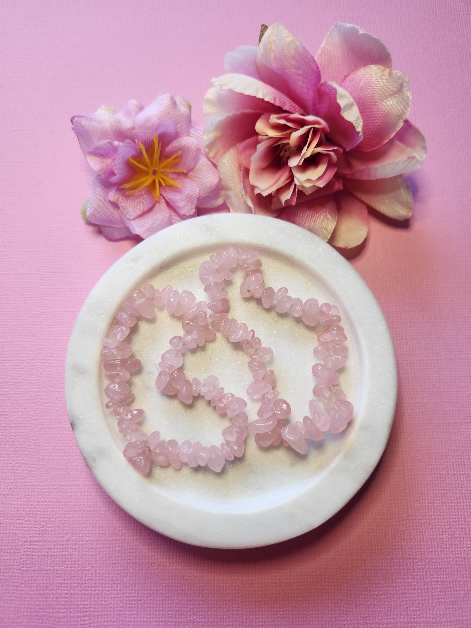 Two Rose Quartz chip bracelets displayed in a white dish with two soft flower props on a light pink background, showcasing their polished pink crystal chips and gentle self-love energy — perfect heart chakra crystal jewelry and a thoughtful healing gift for her.