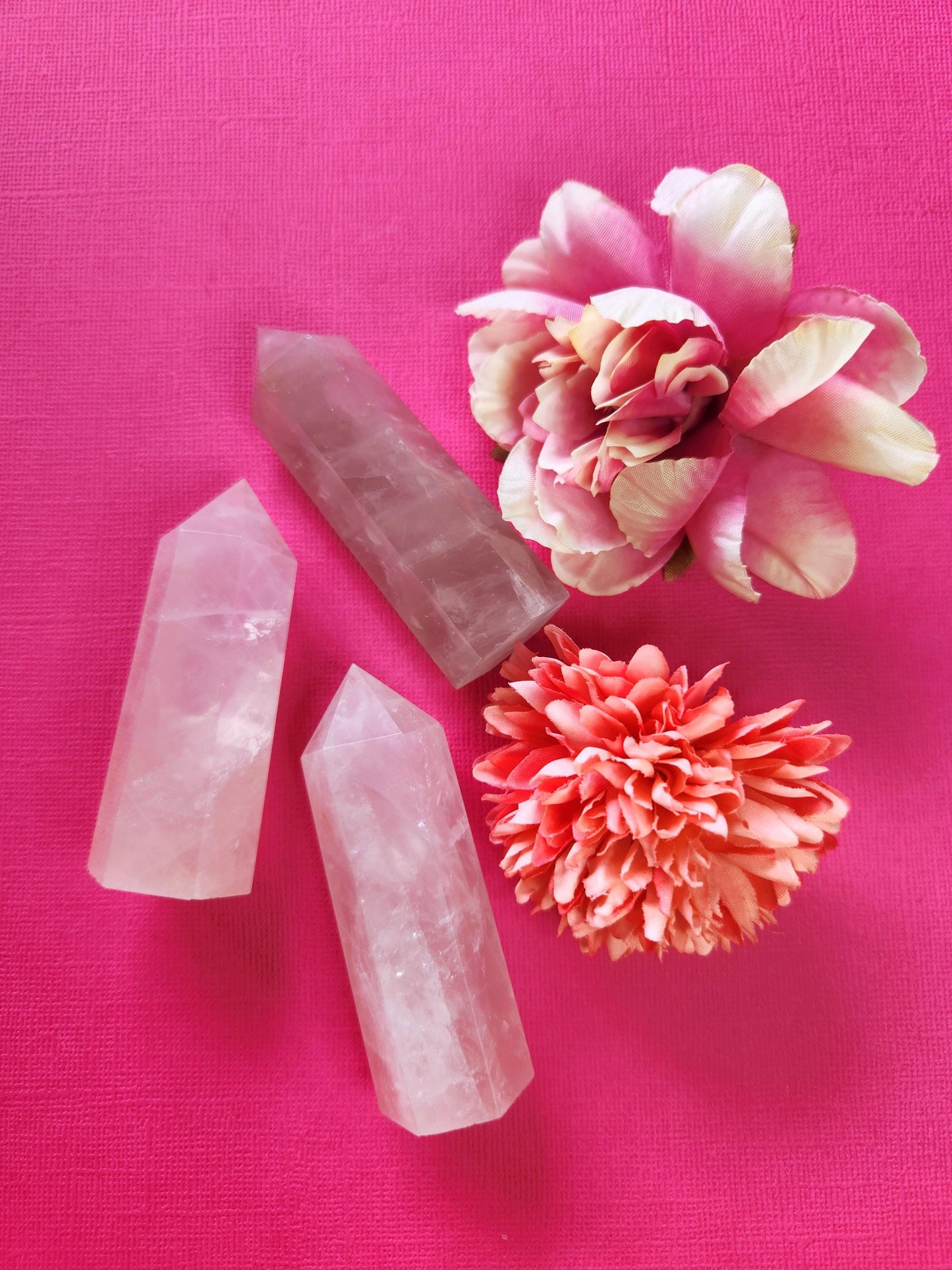 Photo of three polished rose quartz crystal towers standing upright against a bright pink background, accented with two delicate flower props. Their soft pink tones and smooth finish highlight the stone’s loving, heart-healing energy — perfect for crystal decor, self-love rituals, and spiritual balance.