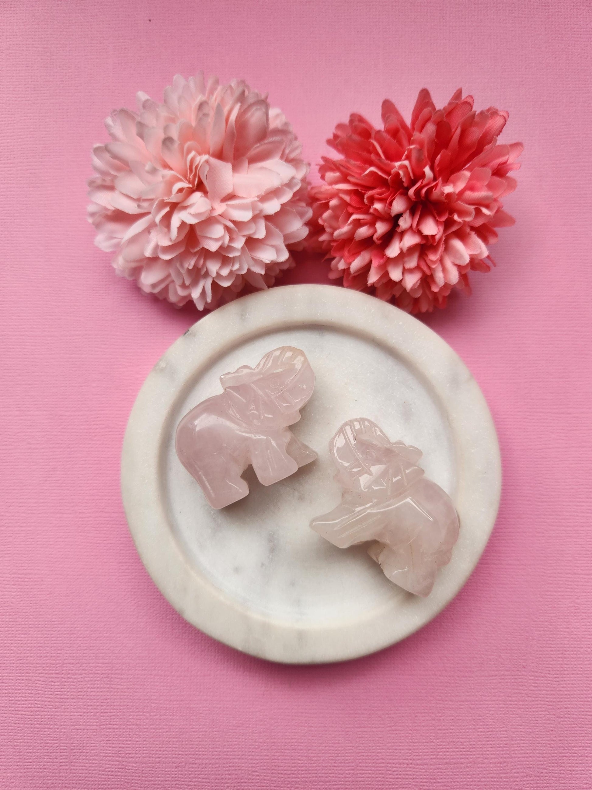 Rose quartz elephant crystal carving displayed in a white dish with floral props on a soft pink background, polished gemstone animal figurine for crystal decor and gifting