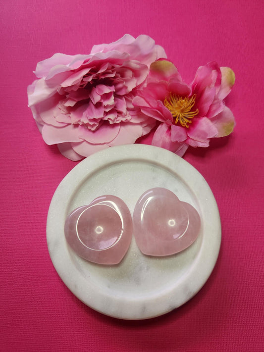 Two Rose Quartz heart-shaped worry stones displayed in a white dish with two flower props on a soft pink background, gentle love crystal pocket stones for self-care and gifting.