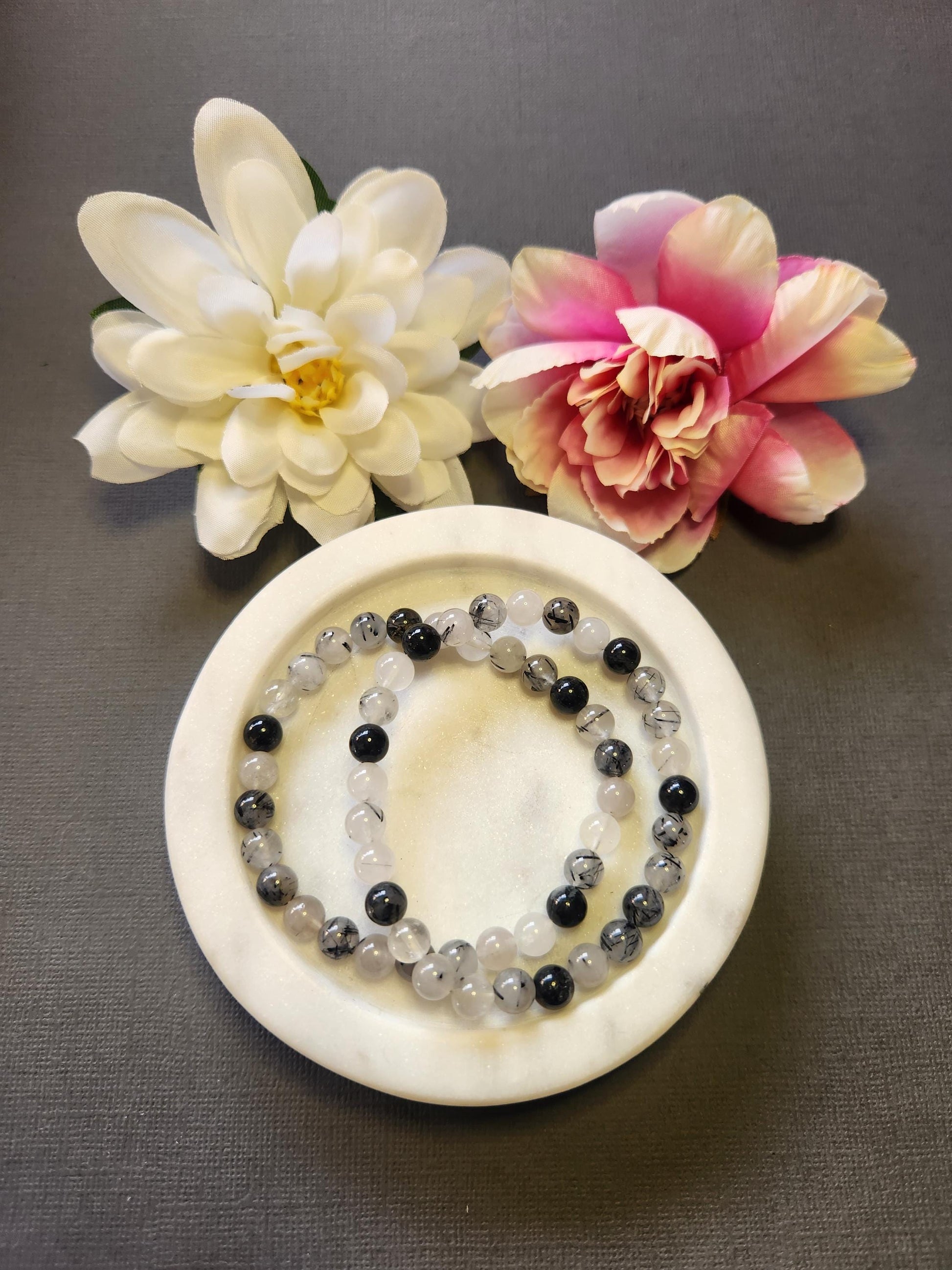 Rutilated Quartz 6mm beaded crystal bracelets displayed in a white dish with two flower props on a dark grey background, featuring natural golden needle-like inclusions in clear quartz beads for energy amplification, clarity, manifestation, and spiritual healing gemstone jewelry photography.