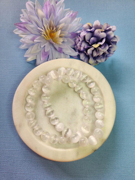 Selenite 8mm beaded bracelets on white dish with two flower props on light blue background, natural selenite crystal beads, handmade cleansing and calming energy bracelet, high-vibration gemstone jewellery product photo