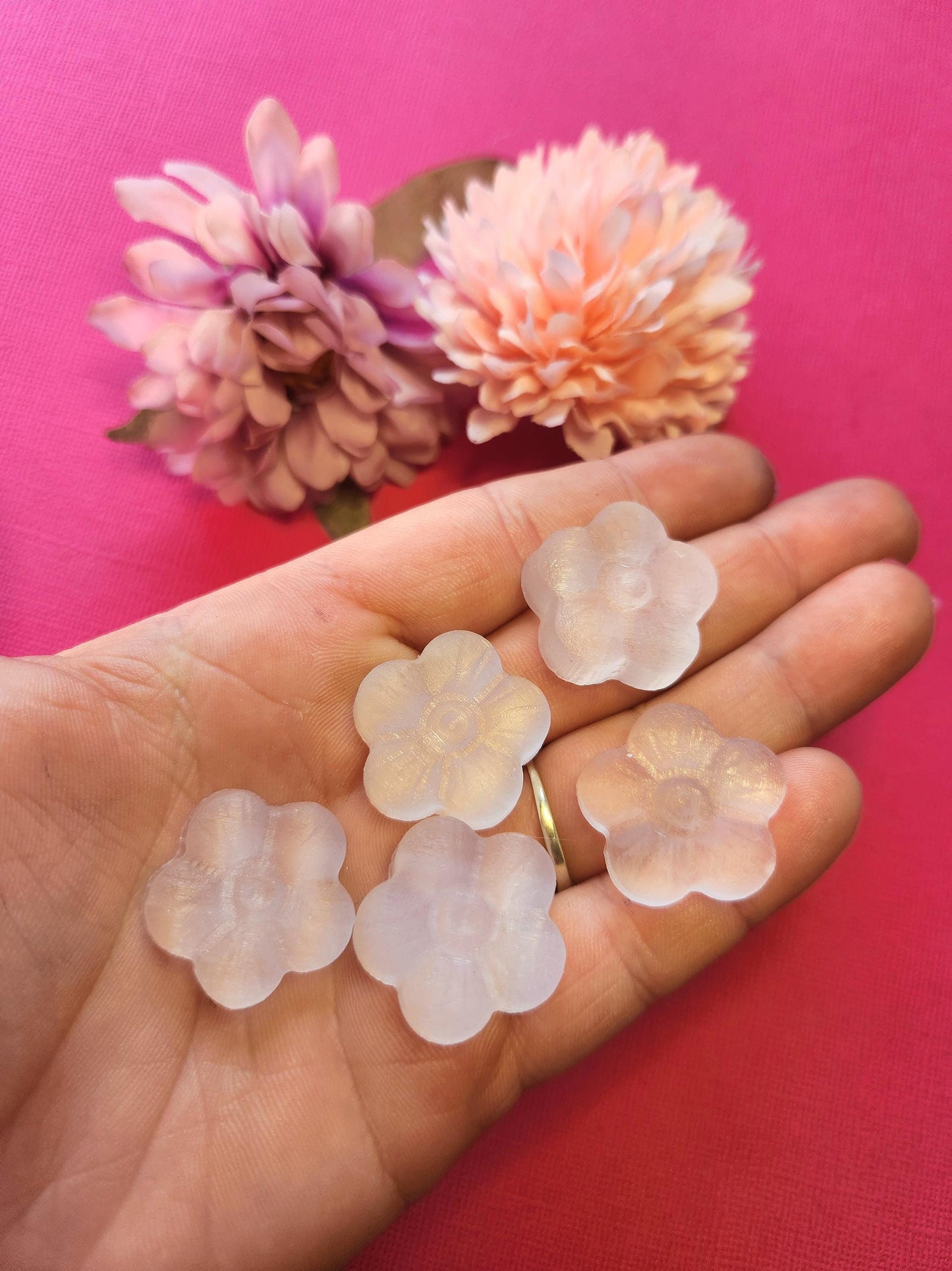 Hand holding natural selenite flower carvings with two pink flower props, set against a bright pink background. Smoothly polished selenite pieces radiate cleansing and calming energy, perfect for crystal collections, meditation, spiritual décor, and gifting.