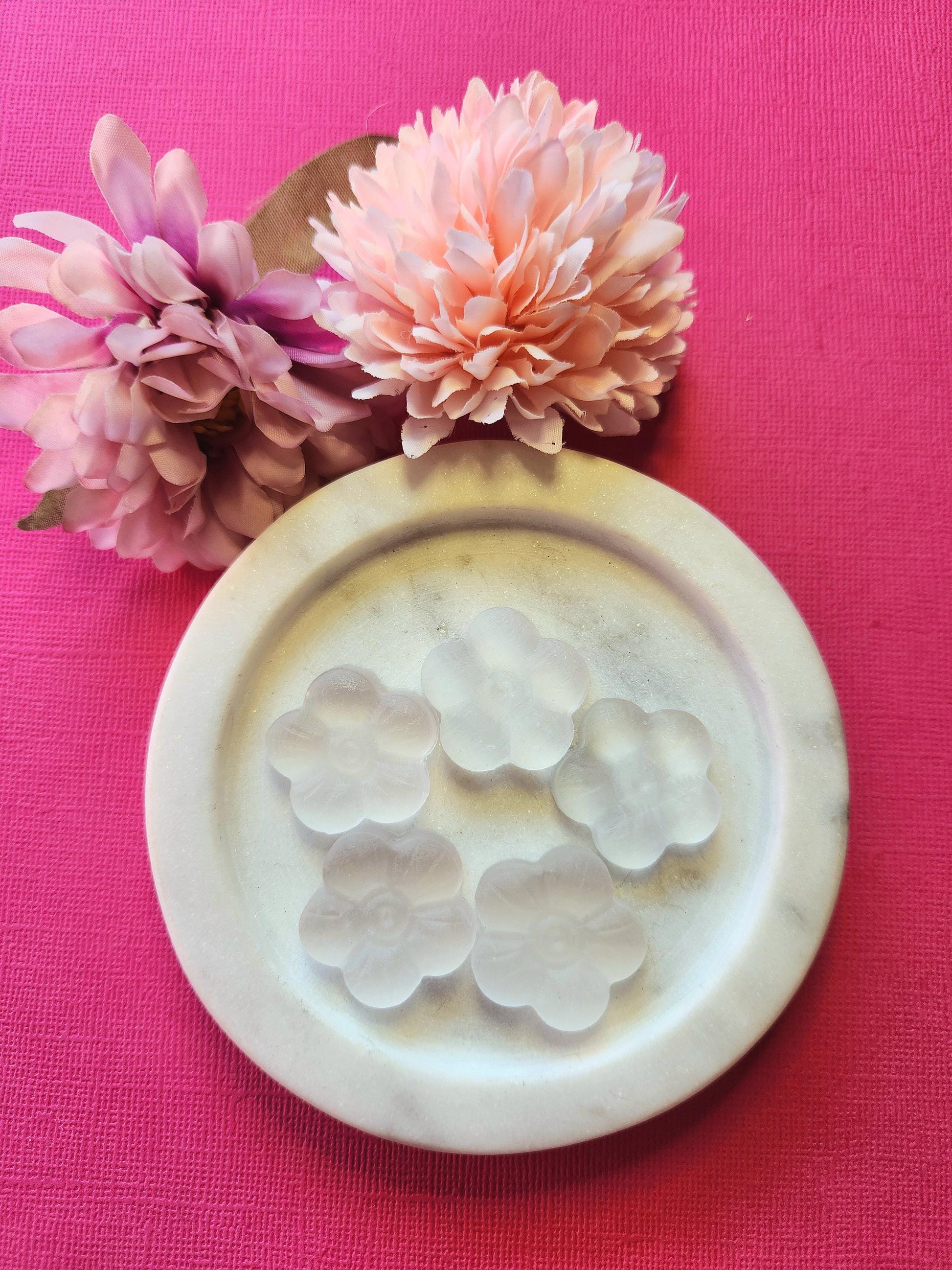 Natural selenite flower carvings displayed in a white dish with two pink flower props, set against a bright pink background. Hand-carved and polished, these selenite flowers promote energy clearing, mental clarity, and spiritual connection — ideal for meditation, crystal collections, and thoughtful gifting.