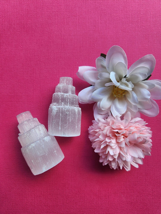 Pair of selenite crystal towers displayed upright on a pink background with delicate flower props featuring polished white selenite points for crystal decor healing meditation and metaphysical energy