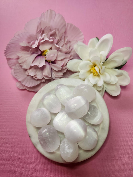 Close-up photo of several selenite tumbles displayed in a white dish with two soft flower props on a pink backdrop. The tumbles show translucent white hues with a smooth, polished finish. Ideal for selenite crystals, tumbled gemstones, crystal collections, and decorative crystal accents