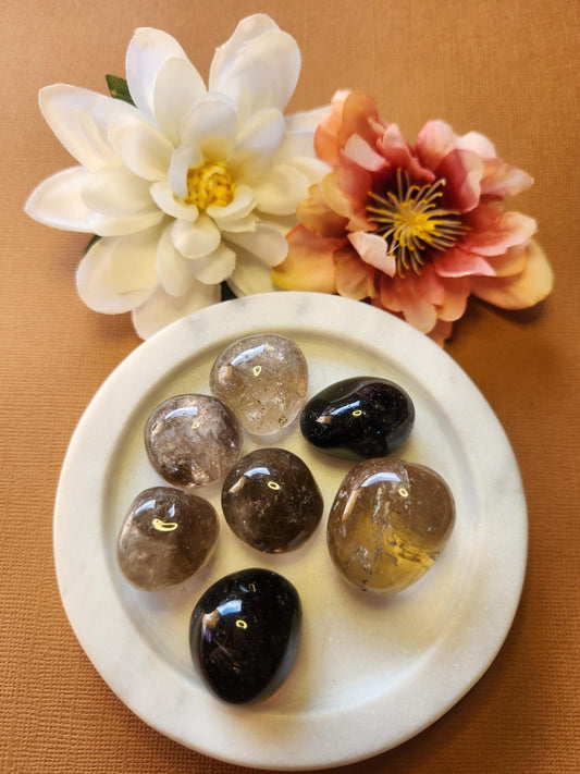 Smoky Quartz tumble stones in a white dish with two flower props on a brown background, polished grounding crystals for protection, stress relief, and chakra healing.