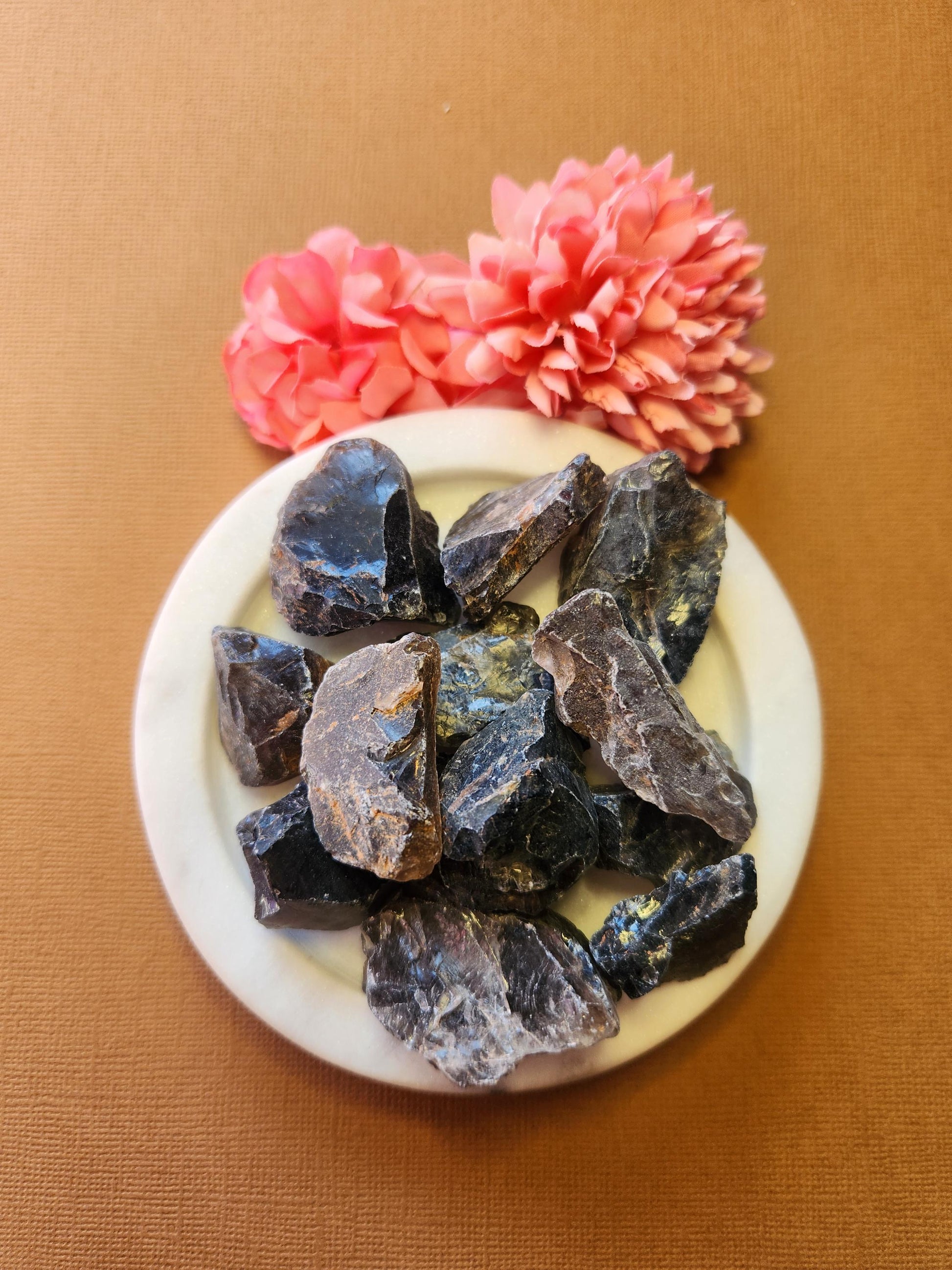 Raw smoky quartz crystal displayed on a white dish with flowers in the background, set against a light brown backdrop, showcasing its natural earthy tones and raw texture.