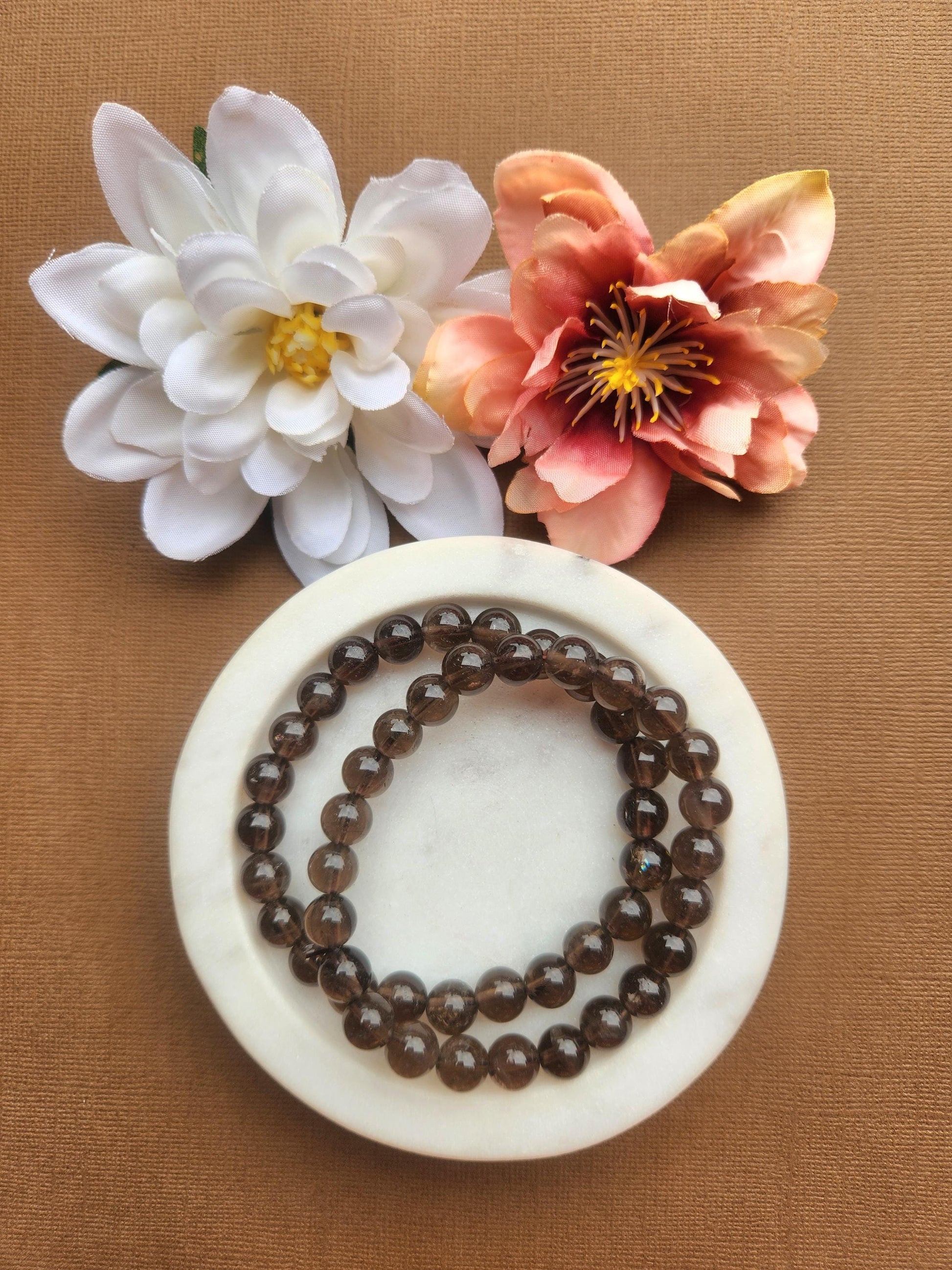 Two smoky quartz 8mm beaded bracelets displayed in a white dish on a brown background with flower props featuring polished brown crystal beads for grounding protection energy cleansing healing jewelry gift