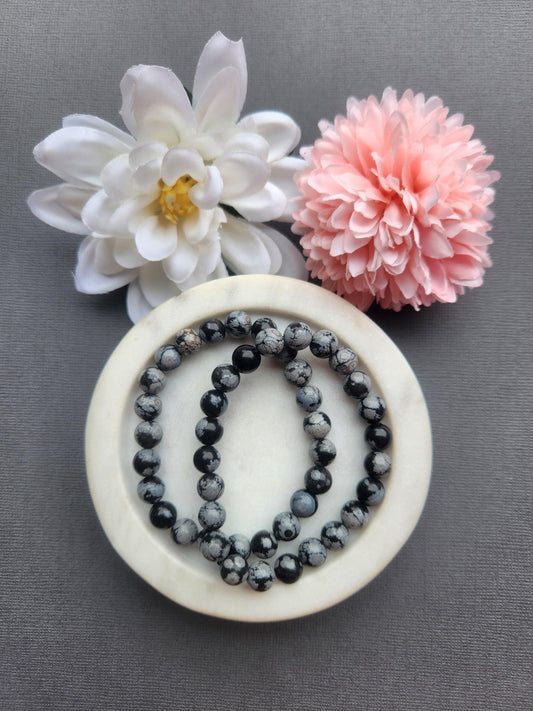Two snowflake obsidian 8mm beaded bracelets displayed in a white dish on a dark grey background with flower props featuring black and white crystal beads for grounding protection balance energy healing jewelry gift