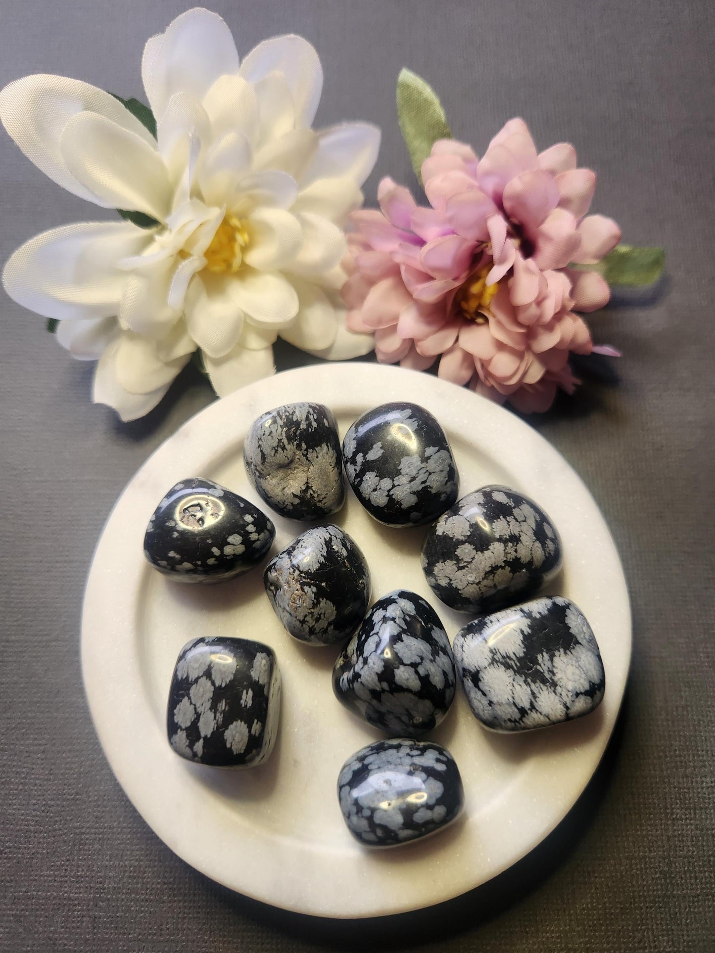 Snowflake obsidian tumble stones displayed in a white dish with two flower props, set against a dark grey background. Polished natural obsidian pieces known for grounding, protection, and balancing energy — perfect for crystal collections, meditation, and spiritual décor.