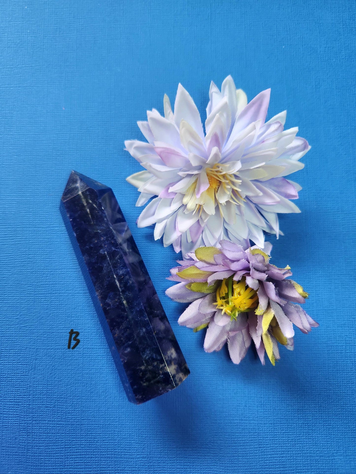 Polished Sodalite Tower B shown with two flower props on a blue backdrop, vibrant blue crystal point with white veining, high-vibration healing stone for intuition, mental clarity, meditation, chakra balancing, manifestation, and spiritual crystal displays