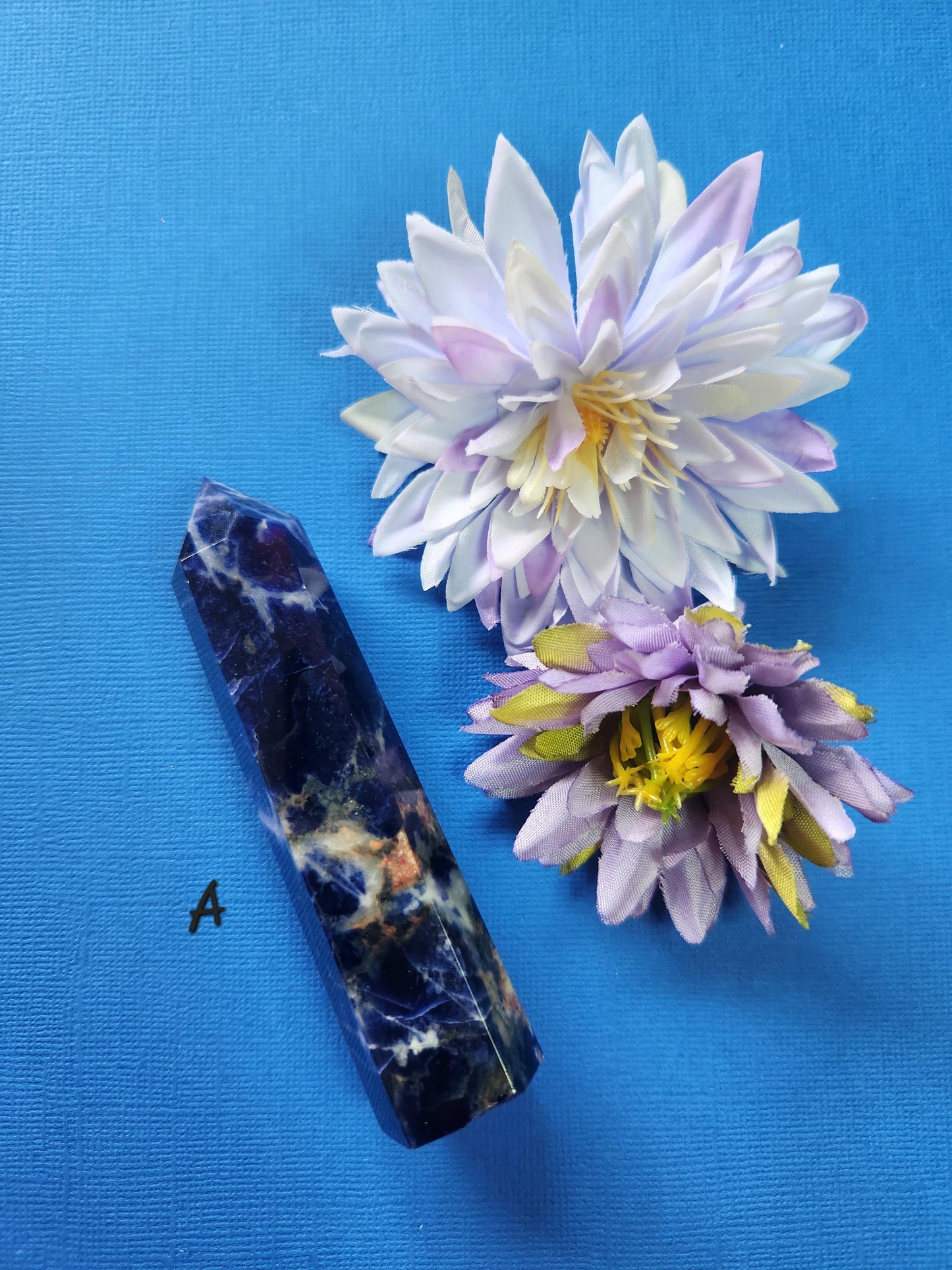 Polished Sodalite Tower A photographed on a blue background with two flower props, deep blue crystal with white streaks, perfect for meditation, psychic development, energy clearing, chakra work, manifestation, spiritual crystal décor, and metaphysical collections.