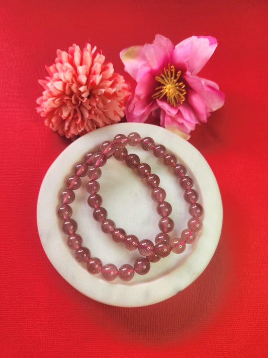 Two Strawberry Quartz 8mm beaded bracelets displayed in a white dish with two flower props on a red background, healing crystal jewellery photo for love energy, heart chakra bracelet image for Etsy and Shopify listings.