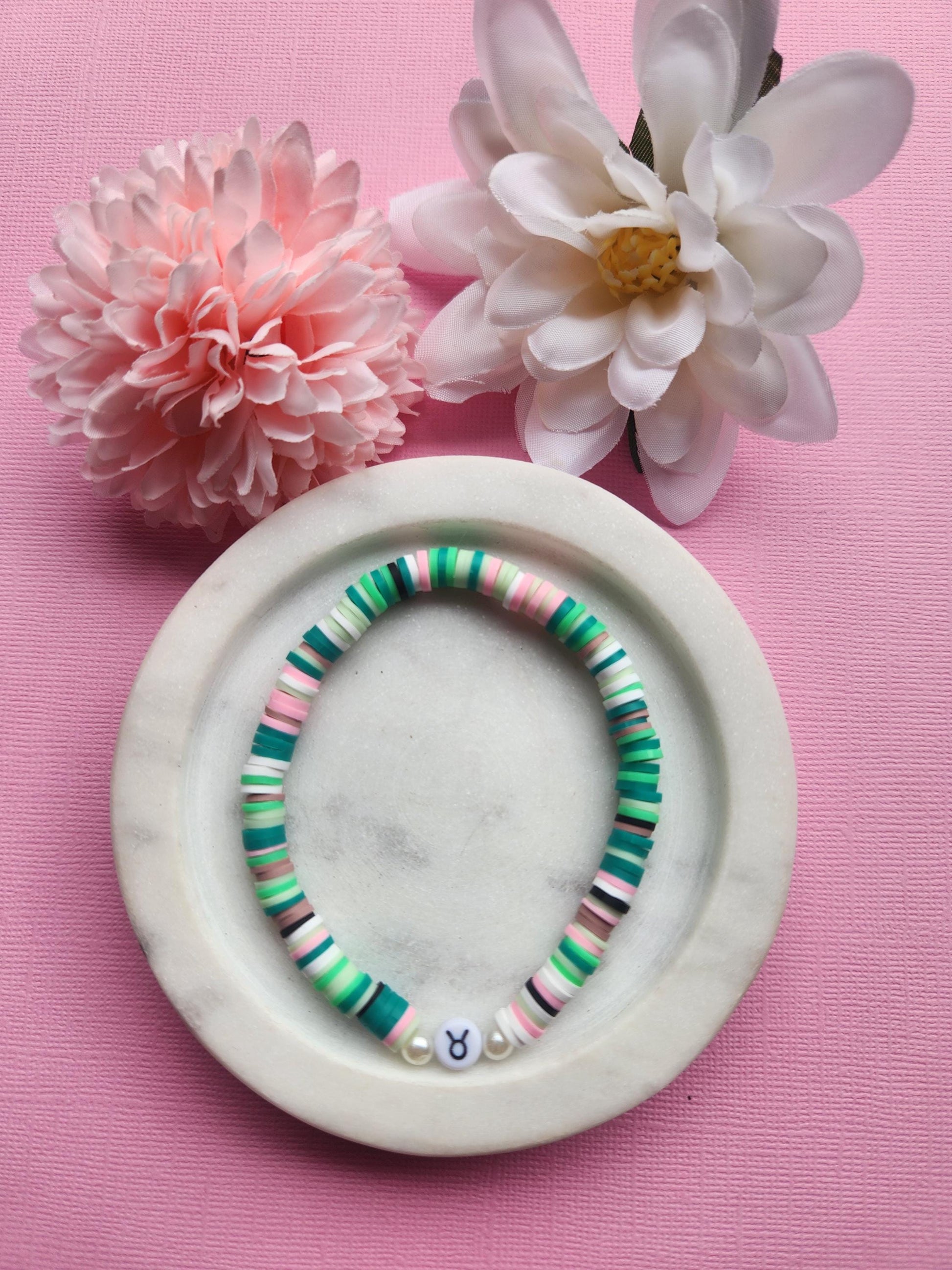 Taurus zodiac clay beaded bracelet displayed in a white dish on a light pink background with two flower props, handmade elastic bracelet featuring pink, green, white, brown and black polymer clay beads with a plastic Taurus symbol bead, astrology jewellery for Taurus gifts, zodiac accessories, handmade beaded bracelets and earthy aesthetic star sign jewellery