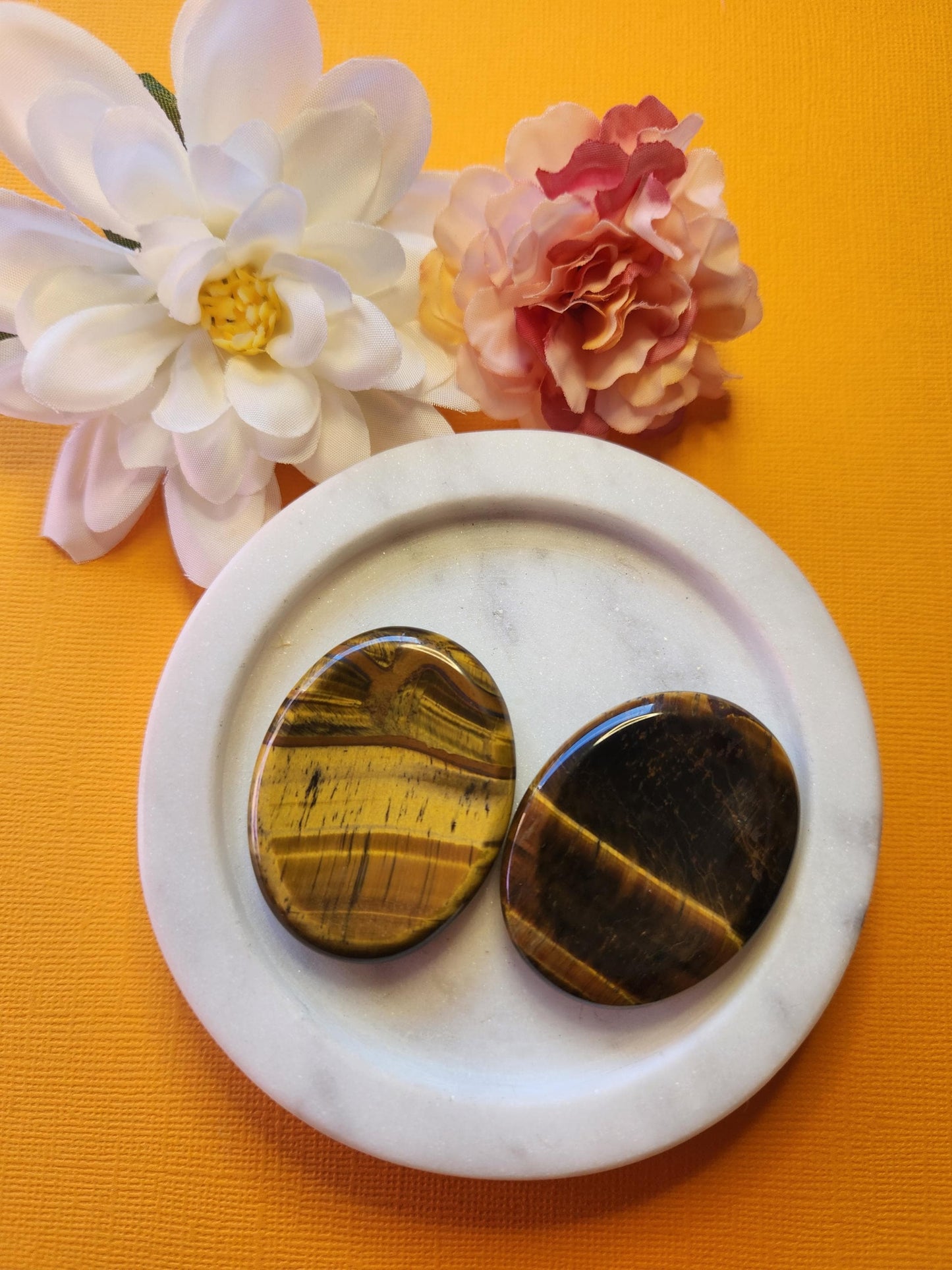 Two polished Tiger’s Eye worry stones on a white dish with two flower props, set against a bright yellow background. Golden-brown thumb stones for courage, grounding energy, and protection.