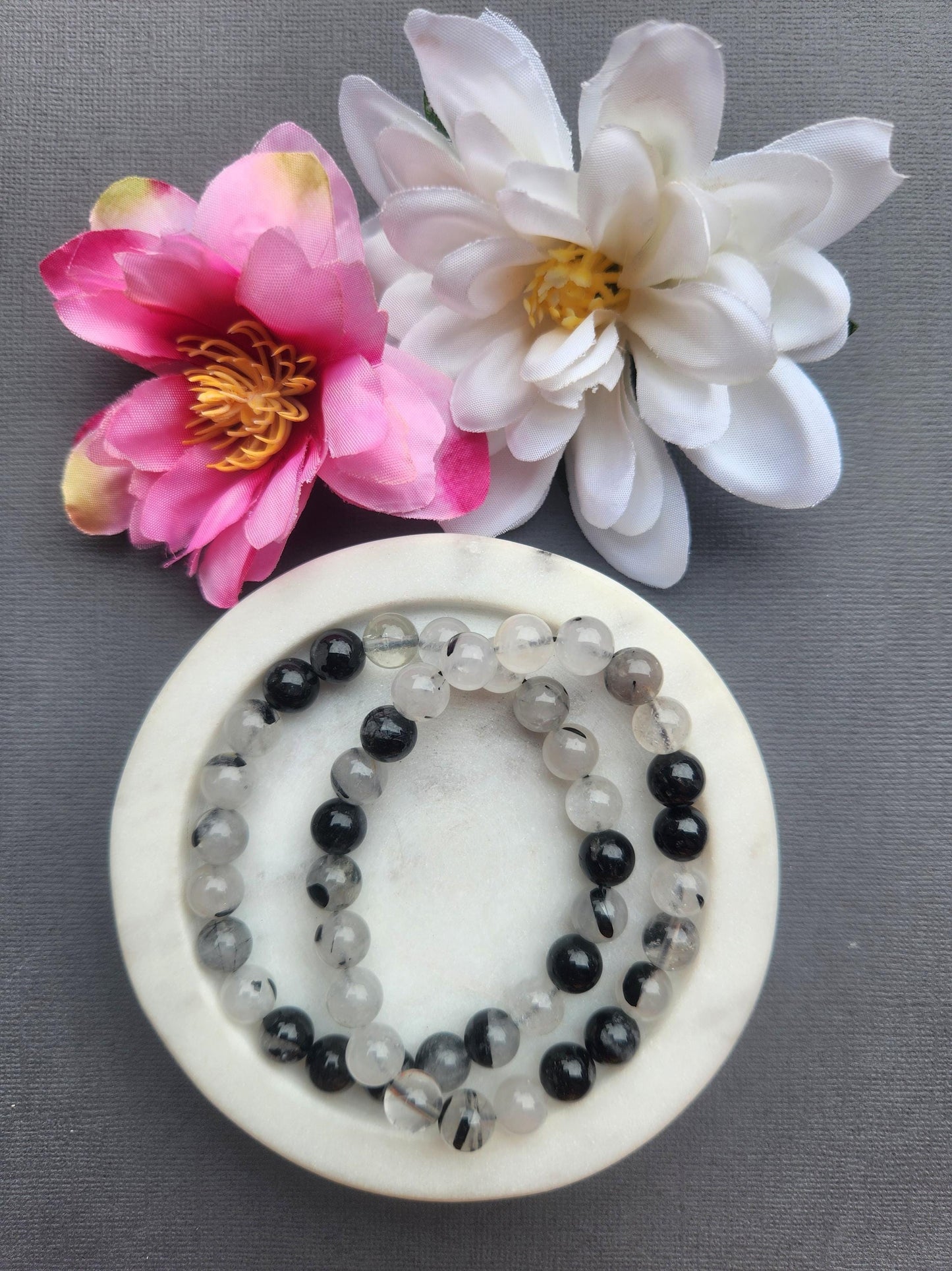Two tourmaline in quartz 8mm beaded bracelets displayed in a white dish on a dark grey background with flower props featuring clear quartz beads with black tourmaline inclusions for protection grounding energy healing jewelry gift
