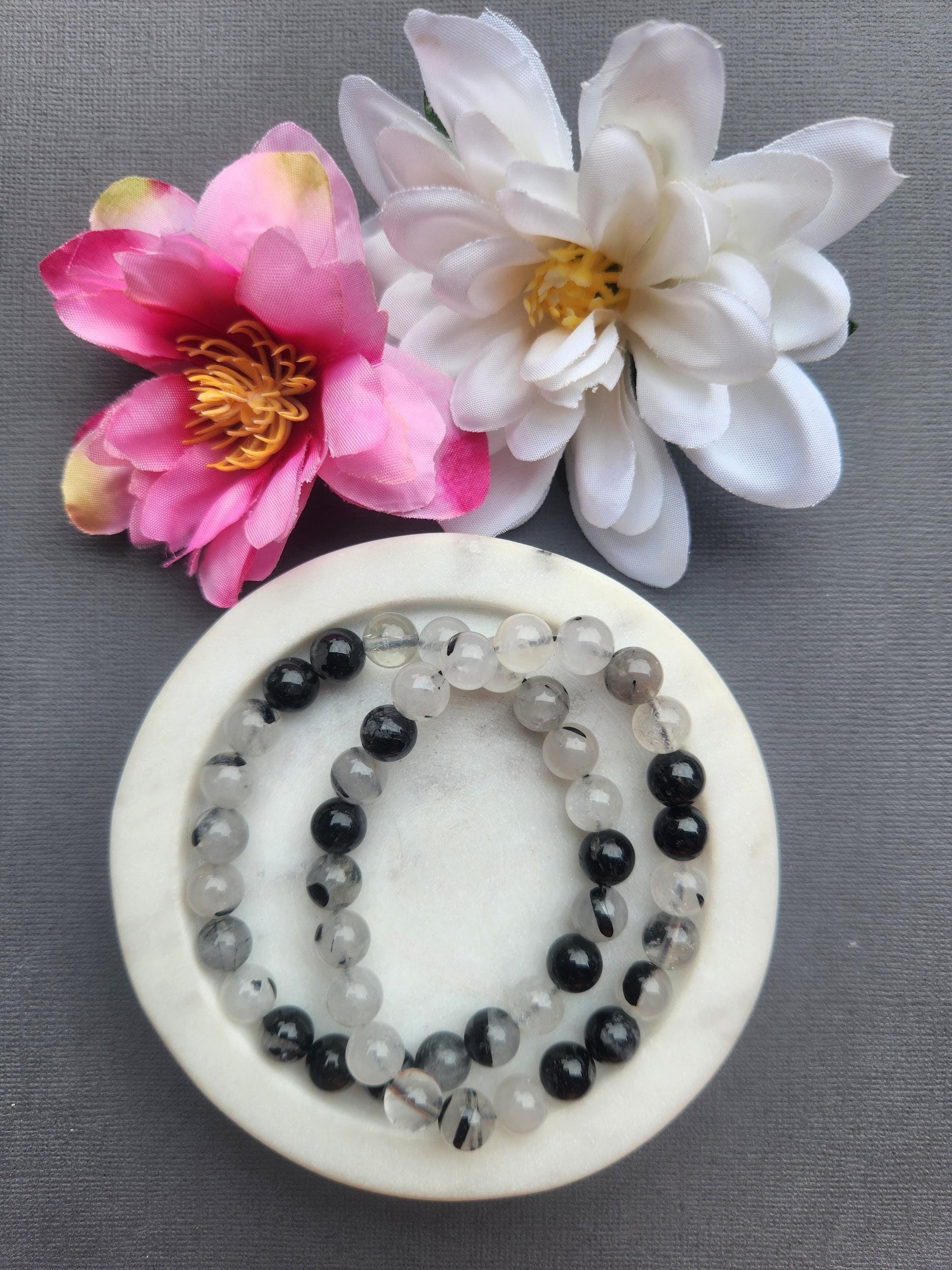 Two tourmaline in quartz 8mm beaded bracelets displayed in a white dish on a dark grey background with flower props featuring clear quartz beads with black tourmaline inclusions for protection grounding energy healing jewelry gift
