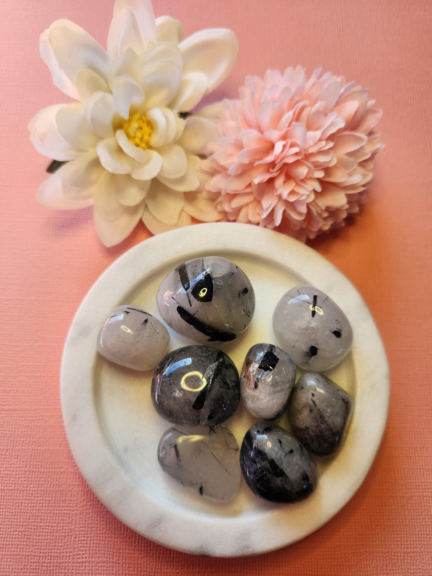 Tourmaline in quartz tumble stones displayed on a white dish with flower props in the background, set against a light pink backdrop, highlighting the unique black needle-like inclusions within clear quartz.
