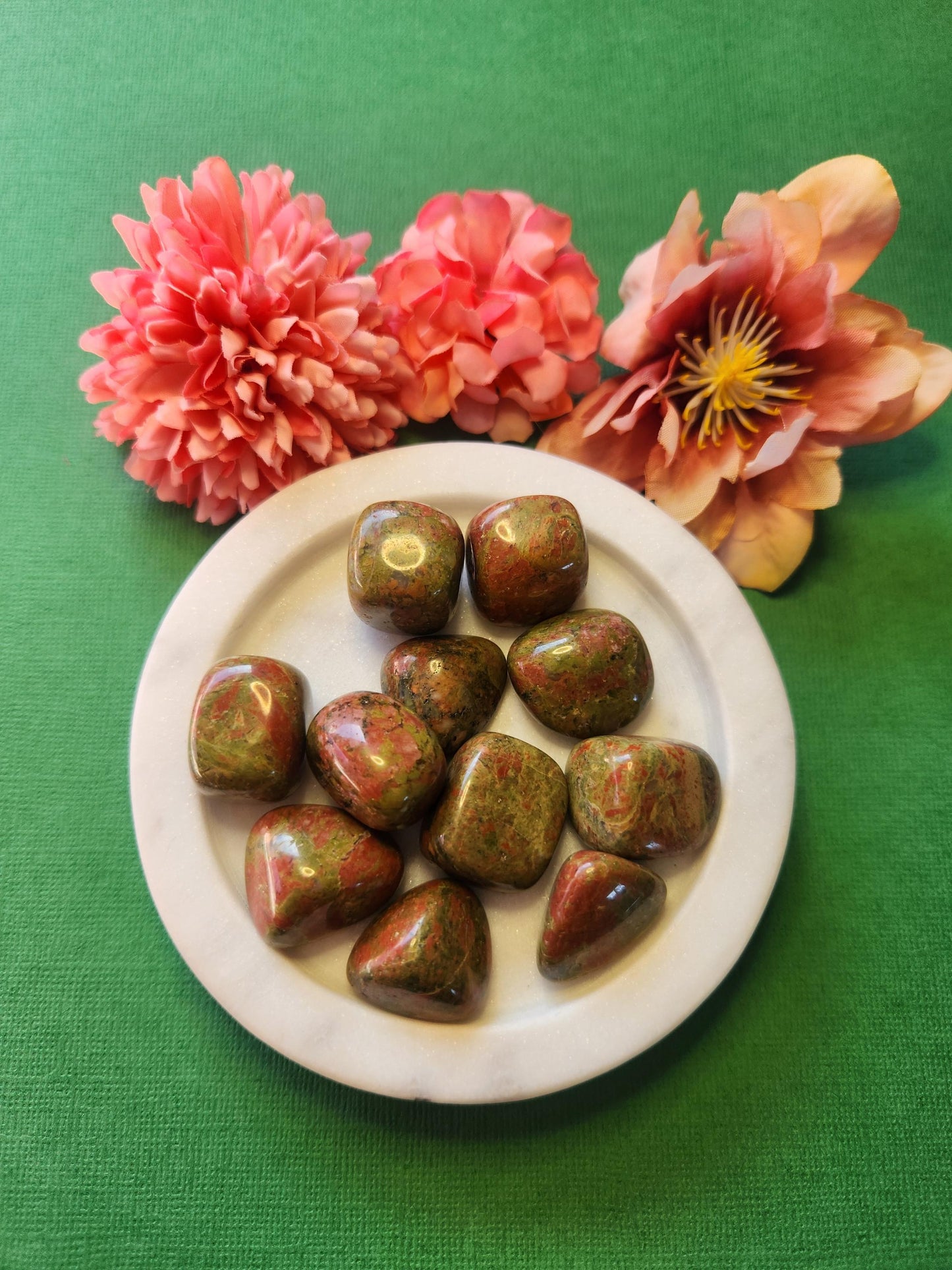 Unakite tumble stones on a white plate, with soft flowers in the background and a green backdrop, creating a natural and earthy display.