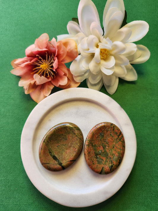 Two polished Unakite worry stones displayed on a white dish, styled with a pink and white flower prop on a green background. Natural green and pink Unakite crystals, perfect for stress relief, emotional balance, meditation, mindfulness, and heart chakra healing. Ideal for crystal collections, reiki, holistic wellness, and calming pocket stones.