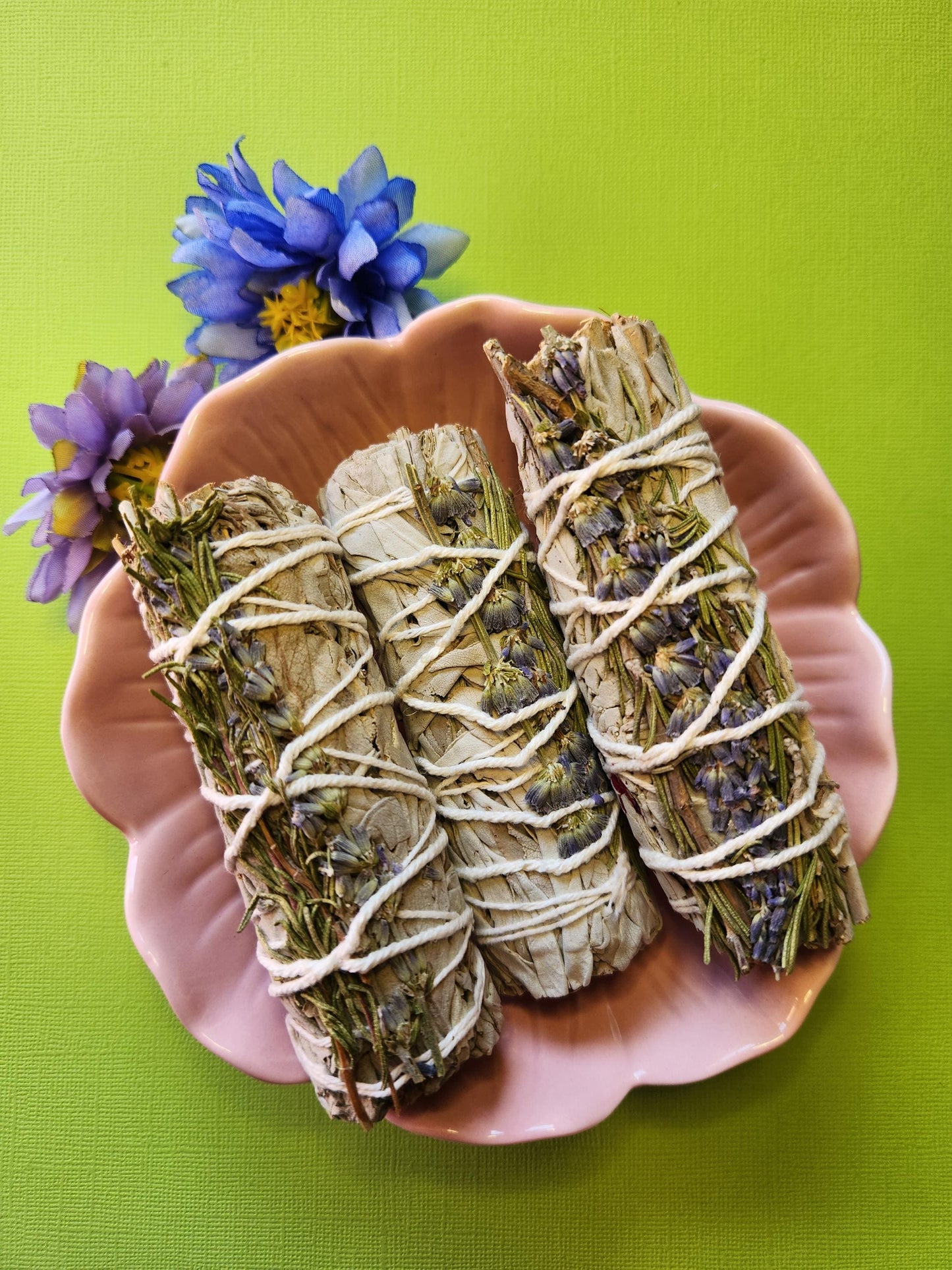 Three White Sage, Rosemary, and Lavender Smudge Sticks displayed in a pink flower-shaped bowl with two flower props on a bright green background — cleansing and calming herbal smudge sticks for energy clearing, relaxation, and spiritual rituals.